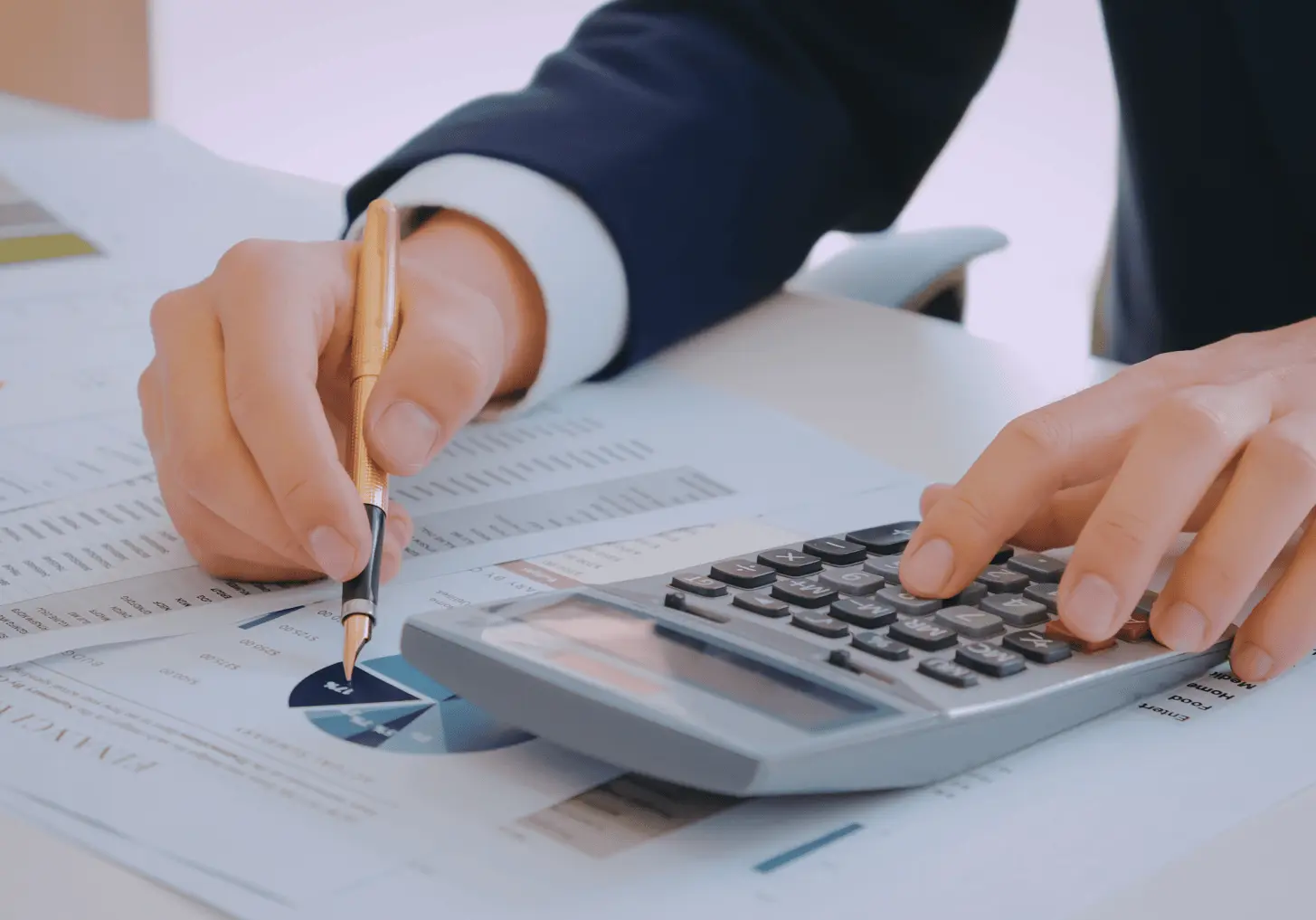 Pessoa de terno analisando relatórios financeiros, utilizando uma calculadora e uma caneta sobre gráficos e tabelas de resultados econômicos.