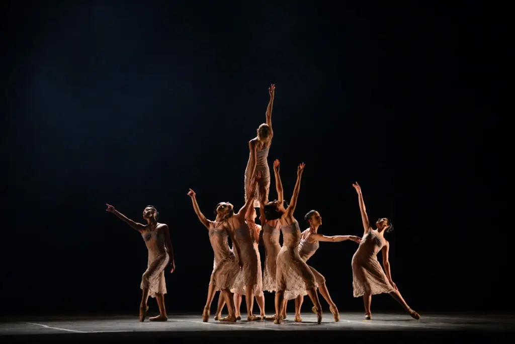 Apresentação de dança em palco escuro no Festival de Dança de Joinville, com grupo de bailarinas em figurinos claros realizando coreografia com movimentos de braços e elevação central.