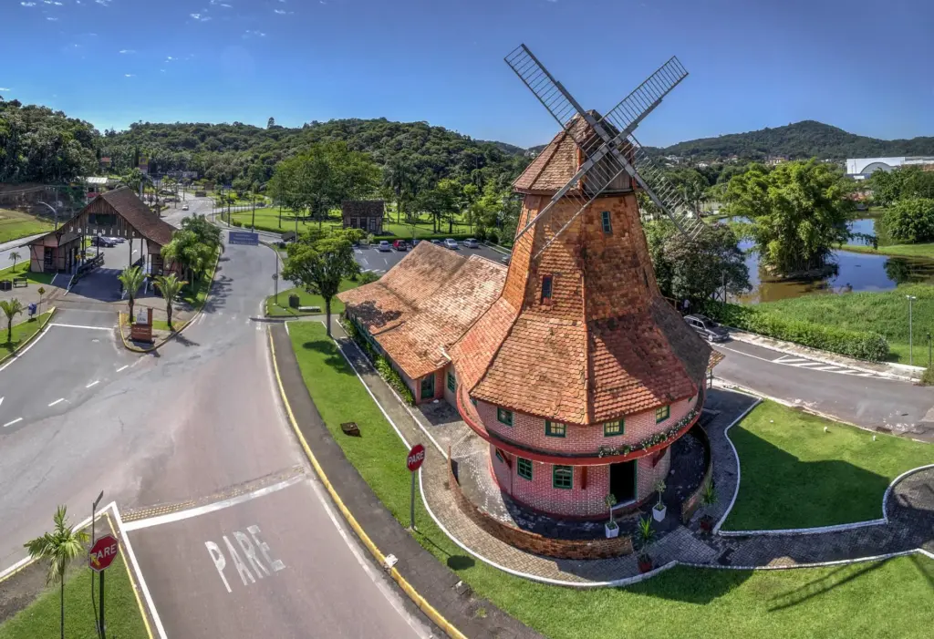 Vista aérea do pórtico turístico e moinho de Joinville, com construção em estilo enxaimel, telhado avermelhado, ruas ao redor, lago, áreas verdes e morros ao fundo.