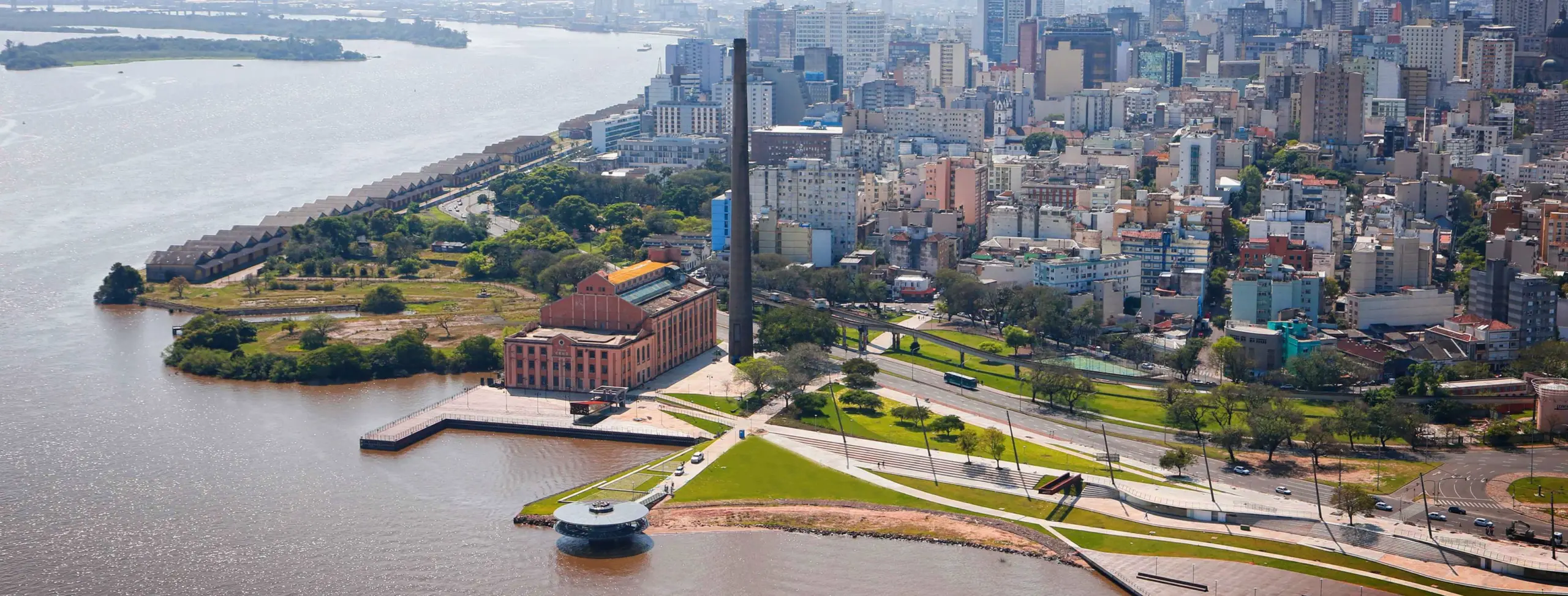 Vista aérea do Cais Mauá, em Porto Alegre, com o Lago Guaíba ao lado, antigos armazéns, a Usina do Gasômetro, áreas verdes e prédios do centro histórico ao fundo.