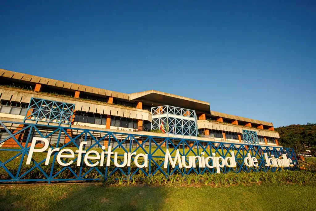 Fachada da Prefeitura Municipal de Joinville, com letreiro em destaque na entrada, estrutura com tijolos aparentes, grades azuis, área verde e céu aberto ao fundo.