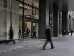 Pessoas em movimento diante da entrada de um edifício corporativo moderno, com fachada de vidro e concreto.