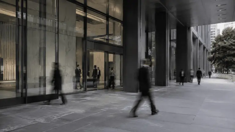 Pessoas em movimento diante da entrada de um edifício corporativo moderno, com fachada de vidro e concreto.