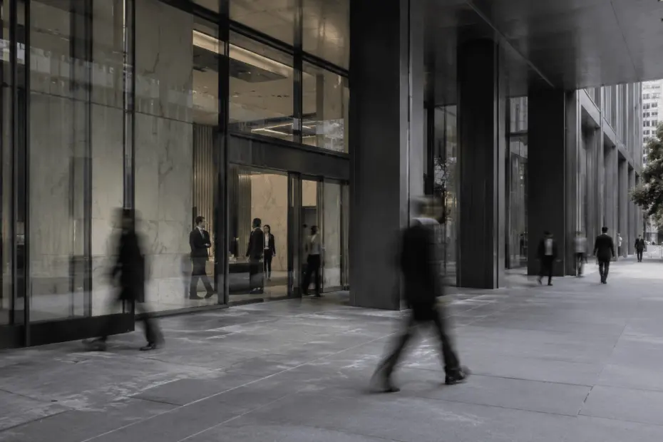 Pessoas em movimento diante da entrada de um edifício corporativo moderno, com fachada de vidro e concreto.