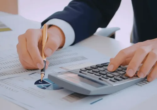 Pessoa de terno analisando relatórios financeiros, utilizando uma calculadora e uma caneta sobre gráficos e tabelas de resultados econômicos.